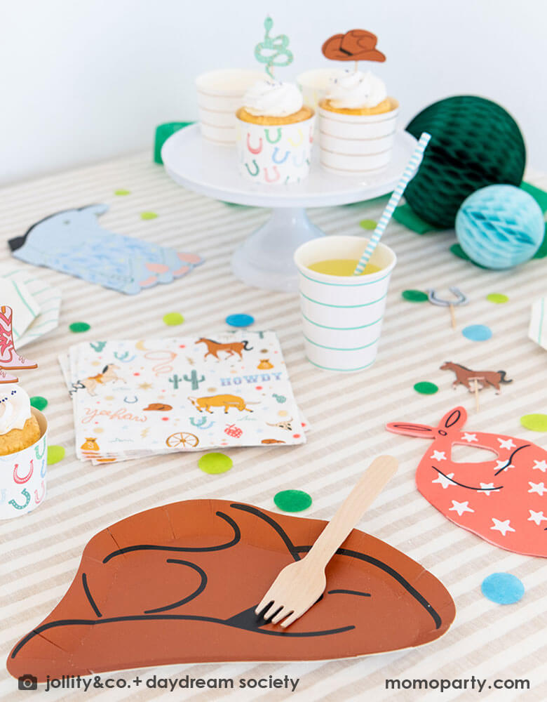 A kid's western themed party table features Momo Party's cowboy themed partyware by Daydream Society including a cowboy shaped plate, a red bandana shaped guest napkin, a Western rodeo themed paper napkin and a blue cowboy boots shaped napkin. On the table there are some cupcakes decorated with cowboy themed toppers, with blue tone honeycomb decorations in back and color coordinated round confetti spread out, this makes a perfect inspiration for a fun rodeo, cowboy themed celebration.