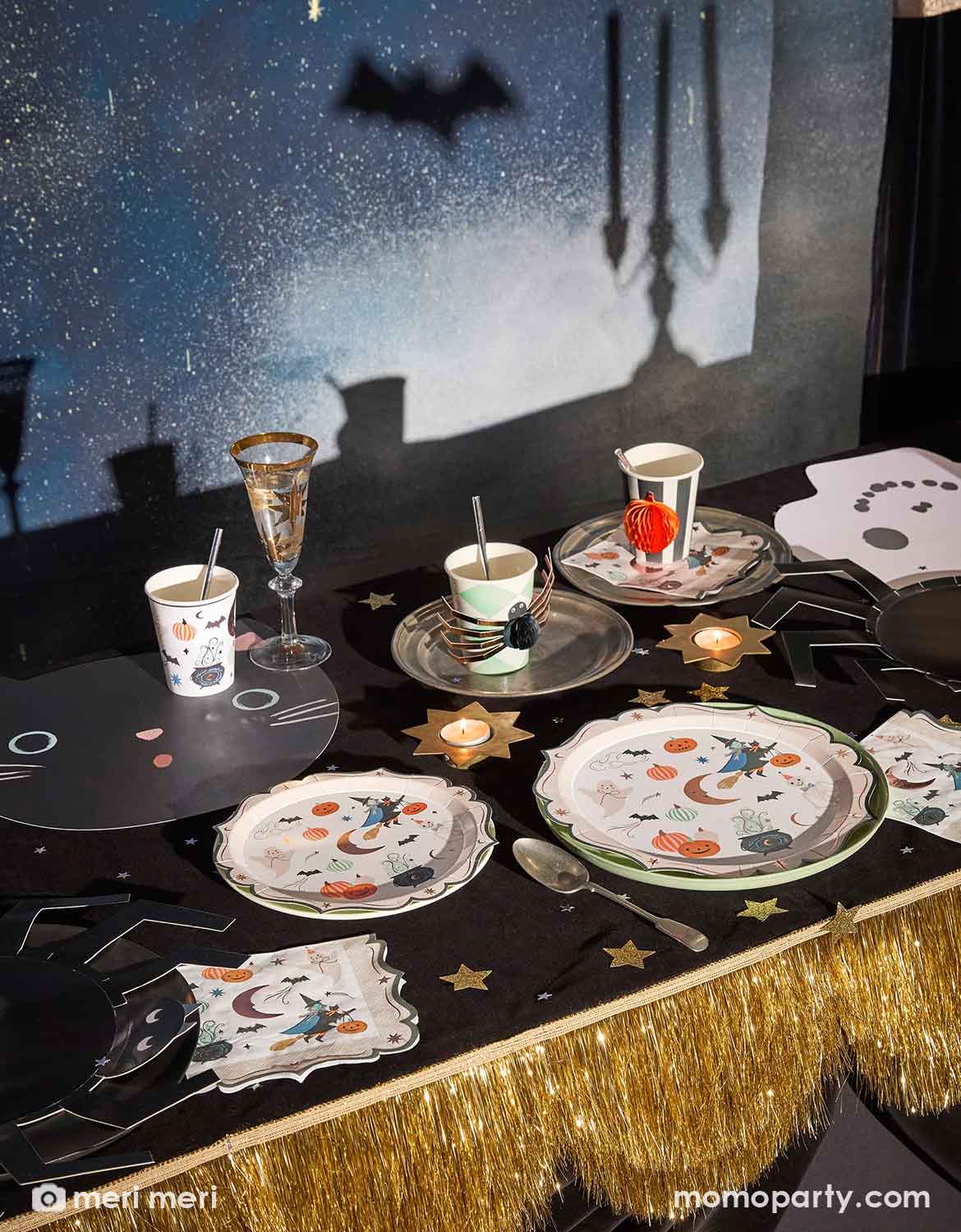 A Halloween-themed party table setting featuring Meri Meri's printed plates, cups, napkins, and a black tablecloth with gold stars and a gold fringe drape from Momo Party. The backdrop is a dark blue starry sky with a bat silhouette and castle shadows.