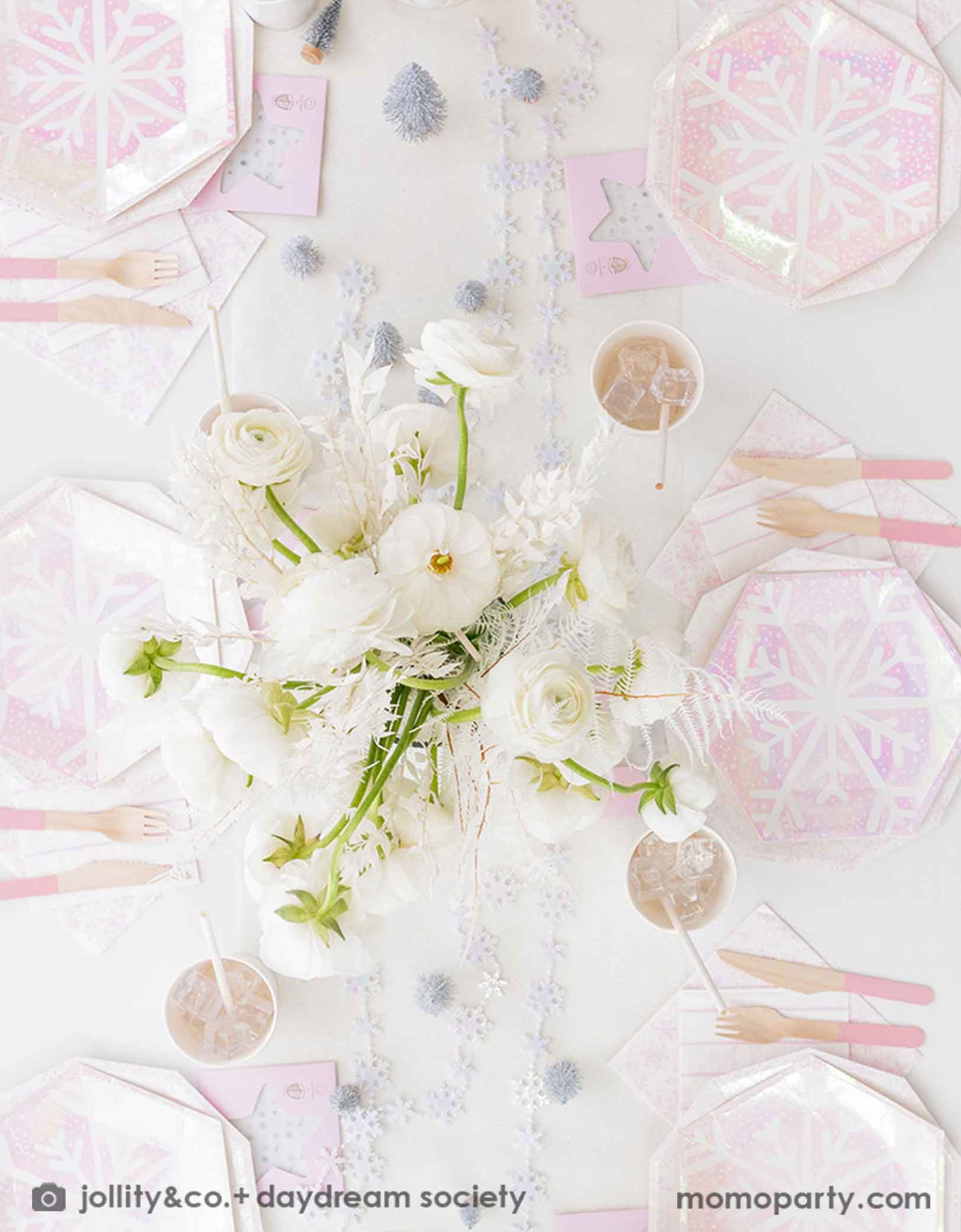 An overhead shot of an elegant Holiday party table setting with white flowers, snowflake themed party tablewares from Momo Party including plates, napkins and party cups. With pink cutlery and decorative Christmas trees and snowflake elements on a white tablecloth, this is a perfect inspiration for a "Let it snow" themed Christmas party, kid's Frozen themed birthday party or a winter inspired gathering in this wonderful season
