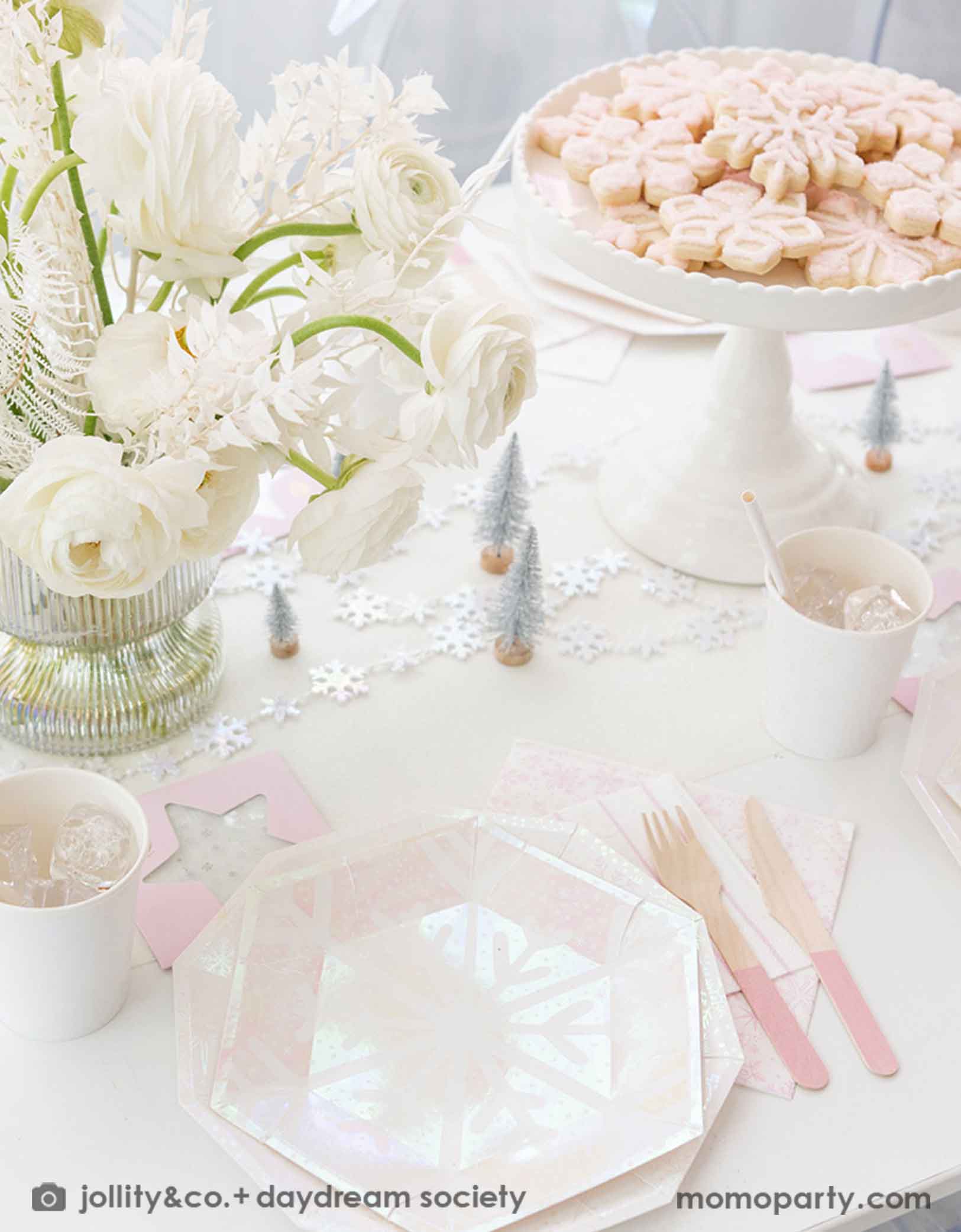 An elegant Holiday party table setting with white flowers, snowflake themed party tablewares from Momo Party including plates, napkins and party cups. With pink cutlery, snowflake shaped cookies on a white cake stand, and decorative Christmas tree elements on a white tablecloth, this is a perfect inspiration for a "Let it snow" themed Christmas party, kid's Frozen themed birthday party or a winter inspired gathering in this wonderful season.