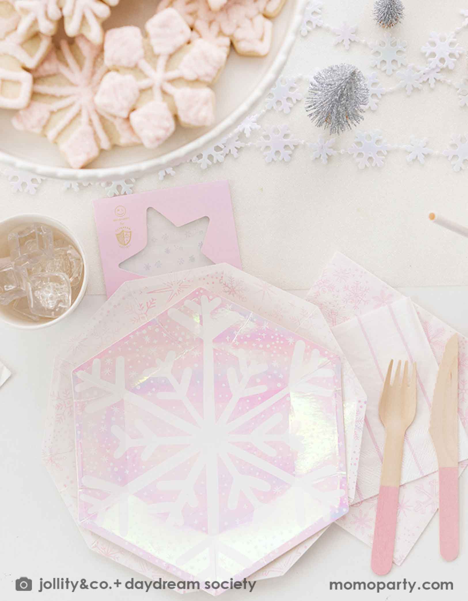 An elegant Holiday party tablescape featuring white flowers, snowflake themed party tablewares from Momo Party including plates, napkins party cups and snowflake temporary tattoos. With pink cutlery, snowflake shaped cookies on a white cake stand, and decorative Christmas trees and snowflake elements on a white tablecloth, this is a perfect inspiration for a "Let it snow" themed Christmas party, kid's Frozen themed birthday party or a winter inspired gathering in this wonderful season.