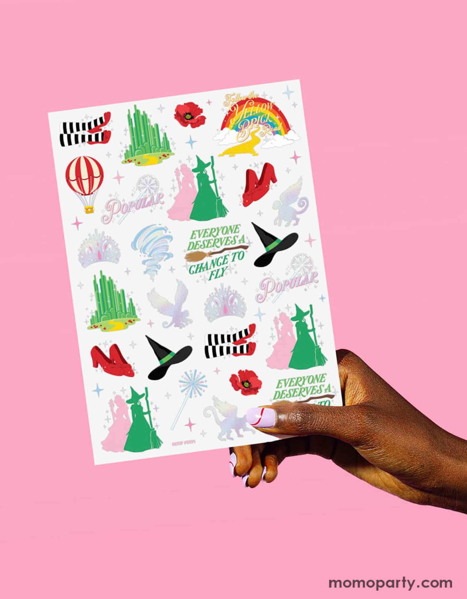 A hand holding Momo Party's Emerald City Wicked Witch Temporary Tattoo sheet by Xo, Fetti. Featuring green and pink witches, witch hats, broomsticks, Emerald city, wands, tiaras and magical icons inspired by the Wizard of Oz. On the table around the tattoo sheets there are some rainbow shaped plates and party cups with pink juice, making it perfect for a Wicked themed birthday party celebration.