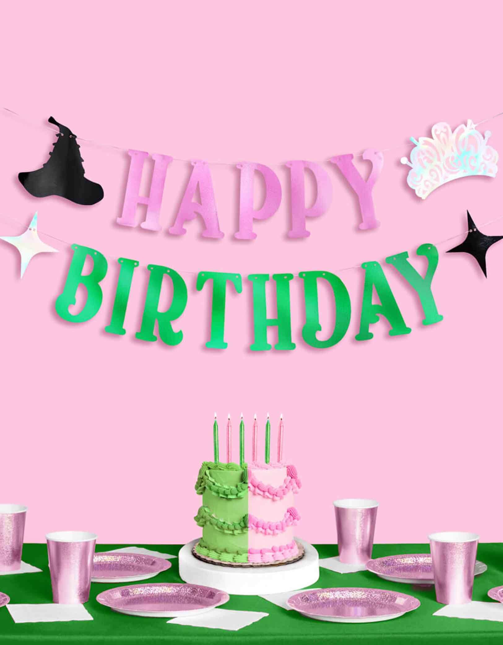 A Wicked themed Birthday party setup with pink and green decorations from Momo Party, including a cake with glittered green and pink birthday candles, the Wicked 'Happy Birthday' banner, green tablecloth and glittered tablewares.