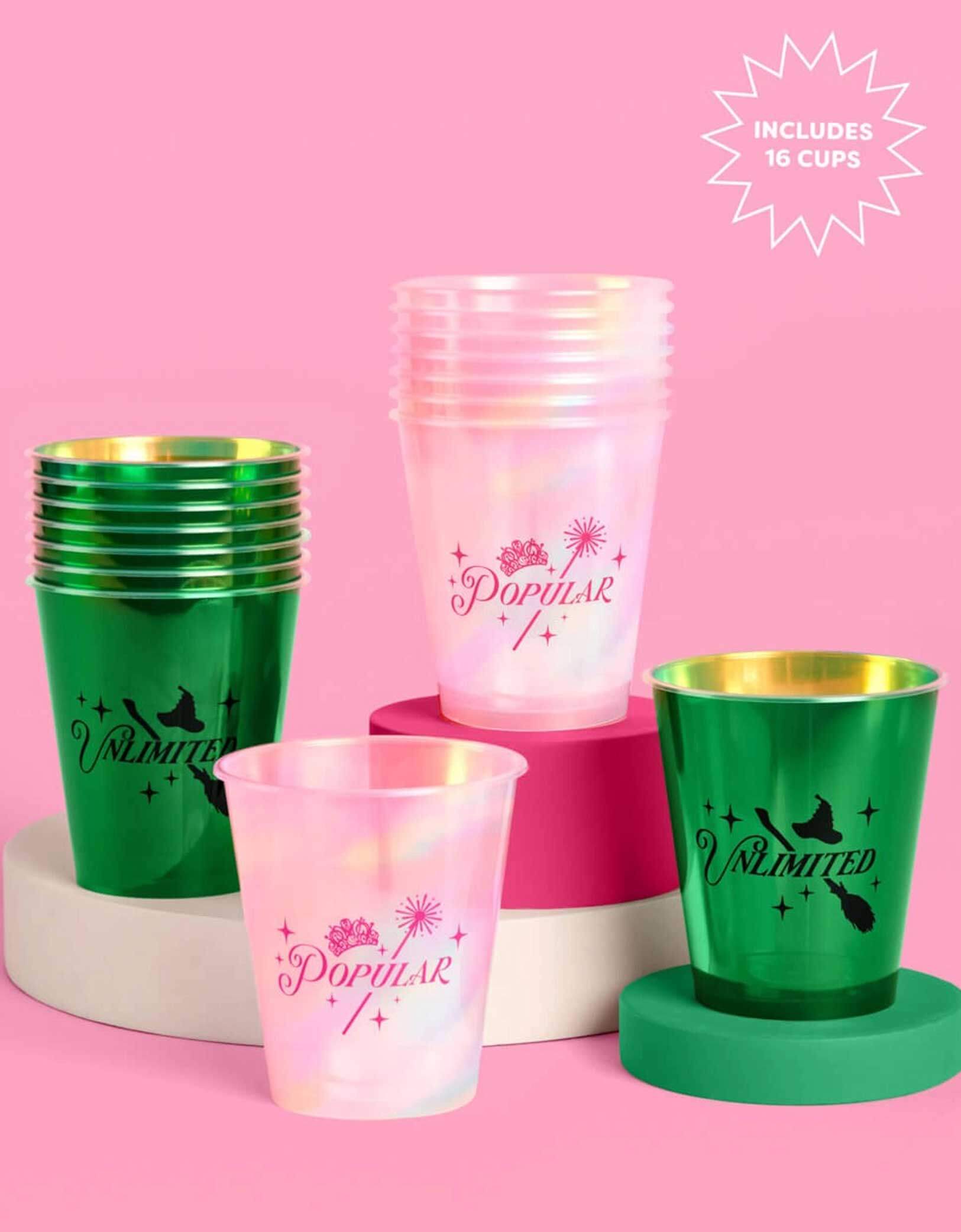A set of Wicked themed party plastic cups from Momo Party, one set of pink cups with "popular" text on it and the other set of green plastic cups with "unlimited" text on it, on a pink platform with a pink background.