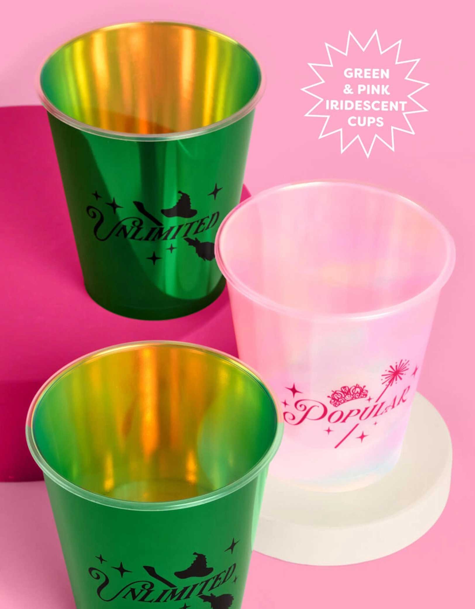 Three Wicked themed party plastic cups from Momo Party, one pink with "popular" text on it and two green cups with "unlimited" text on it, on a pink platform with a pink background.