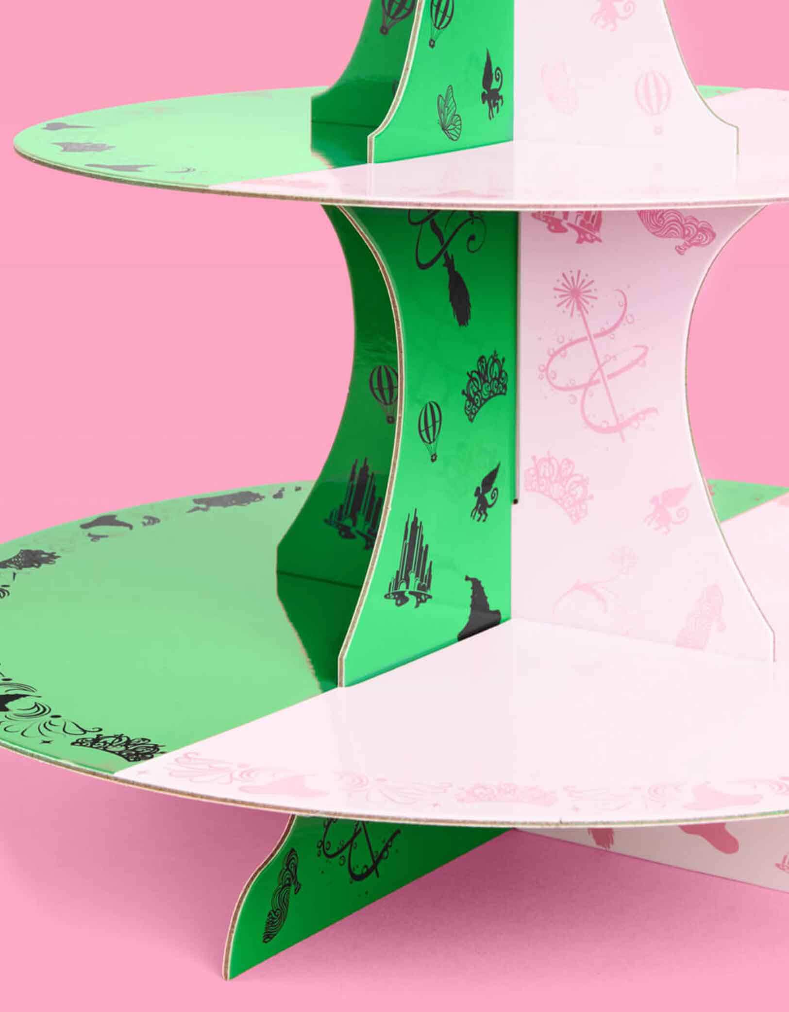 A close up shot of Momo Party's green and pink cake stand with Wicked's Emerald City design at the top on a pink background. Inspired by the enchanting world of the Wicked Witch, this green and pink centerpiece featuring tiaras, wands, hot air balloons, witch hats, and more add a whimsical flair to a Wicked themed celebration. Perfect for showcasing your sweet treats, it transforms your dessert table into a dazzling scene straight from the Emerald City.