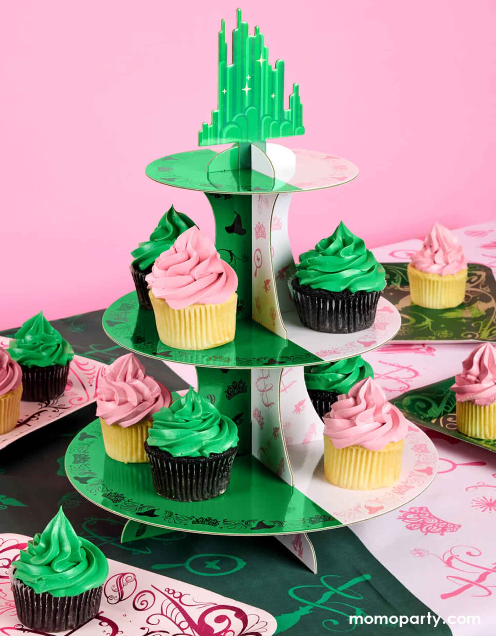 A Wicked themed party table featuring various Wicked themed party supplies from Momo Party including green and pink cupcakes on the Emerald City Wicked shaped cupcake stand, Wicked pink and green tablecloth, and wicked themed party plates against a pink background.