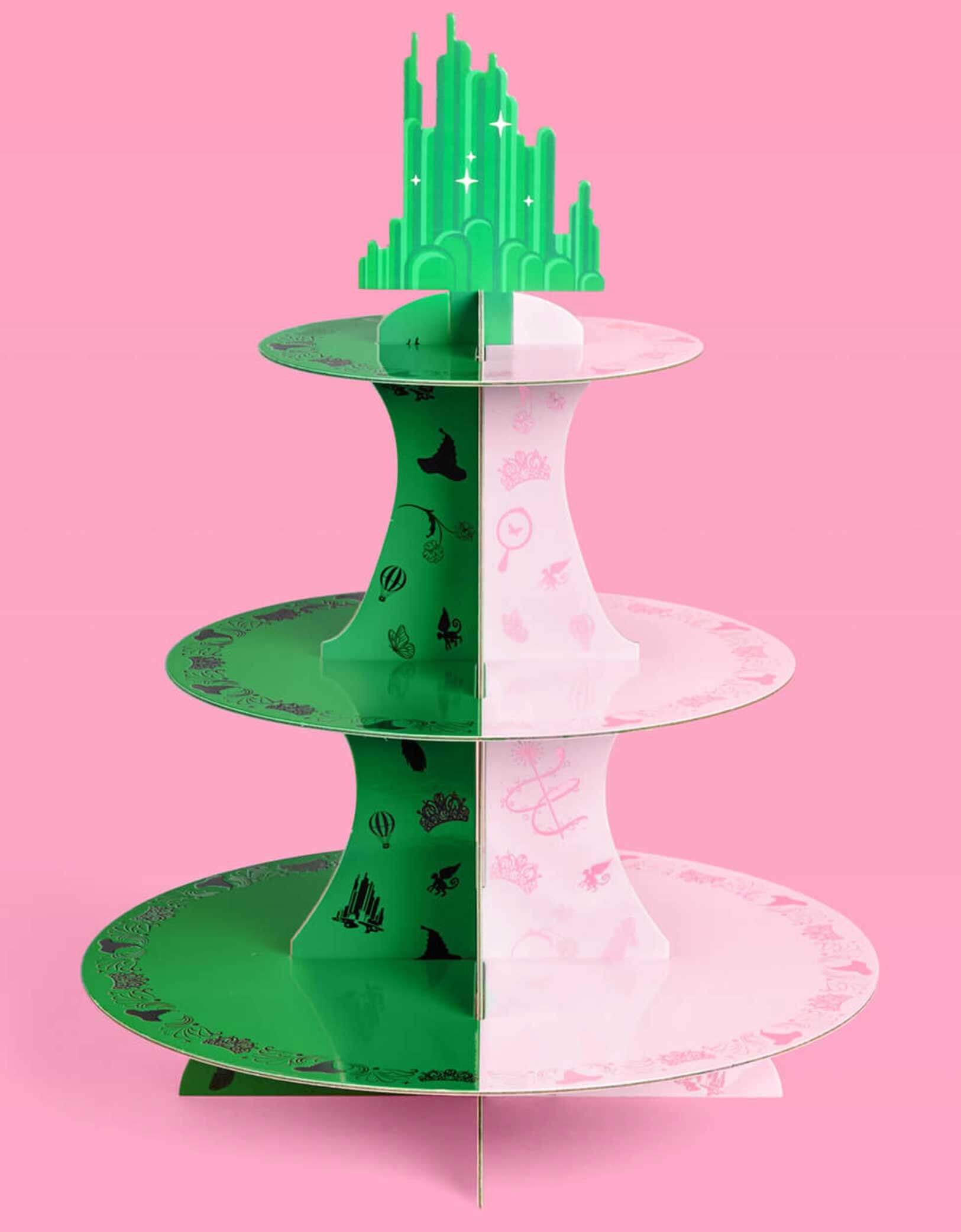 Momo Party's green and pink cake stand with Wicked's Emerald City design at the top on a pink background. Inspired by the enchanting world of the Wicked Witch, this green and pink centerpiece adds a whimsical flair to any celebration. Perfect for showcasing your sweet treats, it transforms your dessert table into a dazzling scene straight from the Emerald City. Topped with a green cityscape with iridescent foil details, let your cupcakes shine and your guests be charmed by this playful, magical displ