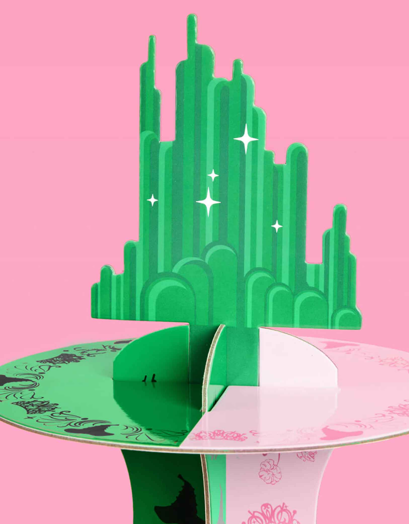 A close up of the Emerald City Topper on Momo Party's green and pink Wicked cake stand on a pink background. Inspired by the enchanting world of the Wicked Witch, this green and pink centerpiece adds a whimsical flair to any celebration. Perfect for showcasing your sweet treats, it transforms your dessert table into a dazzling scene straight from the Emerald City.
