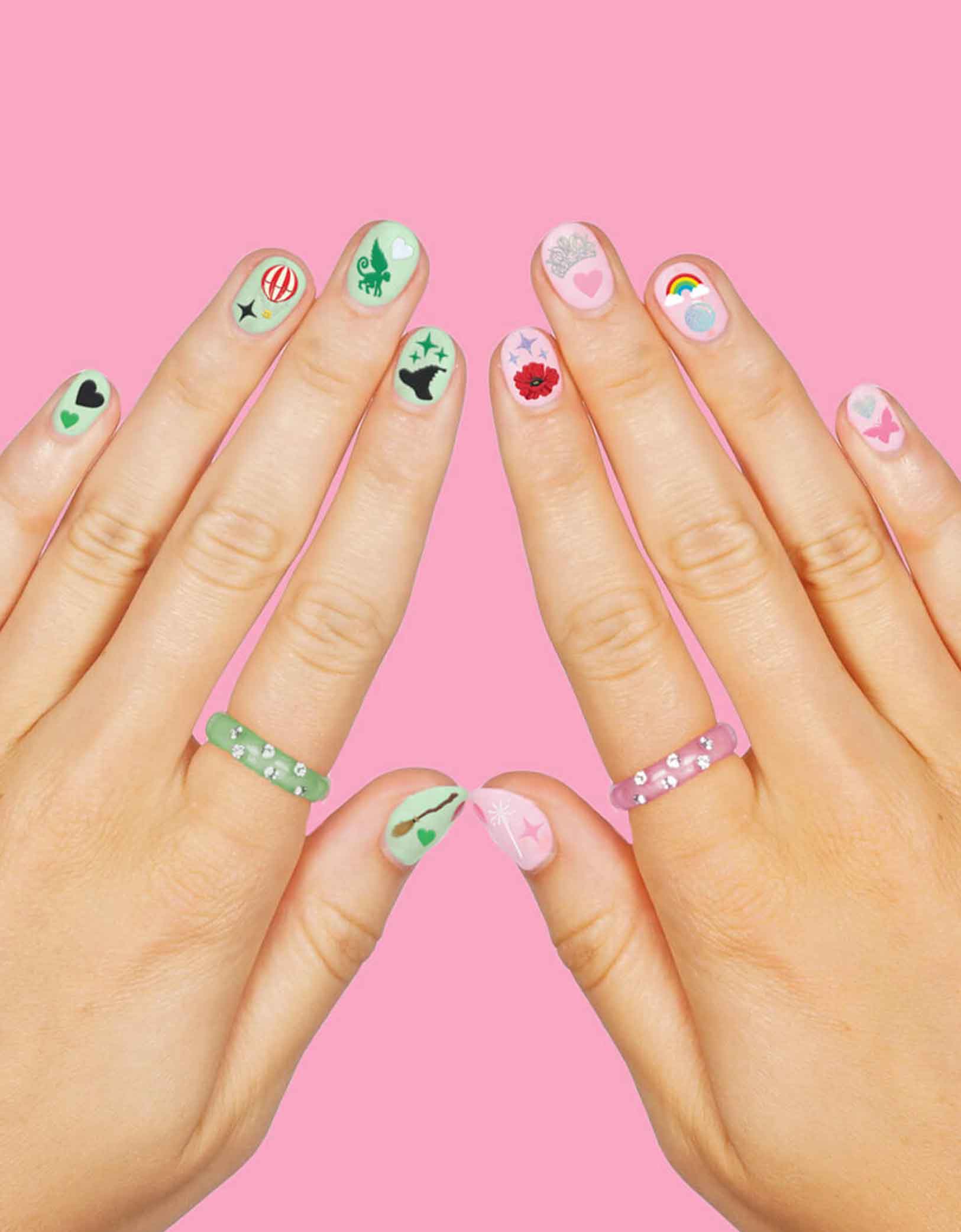 Two hands with with Momo Party's Wicked Emerald City Nail Stickers by Xo, Fetti on a pink background, featuring wicked themed elements including witch hats, bubbles, hot air balloons, butterflies, tiaras, rainbows, sparkles, witch's brooms, they’re perfect for a fun, themed activity that lets guests express their playful side. Easy to apply and remove, these stickers bring a splash of fantasy to your fingertips, making every celebration a little more spellbinding.