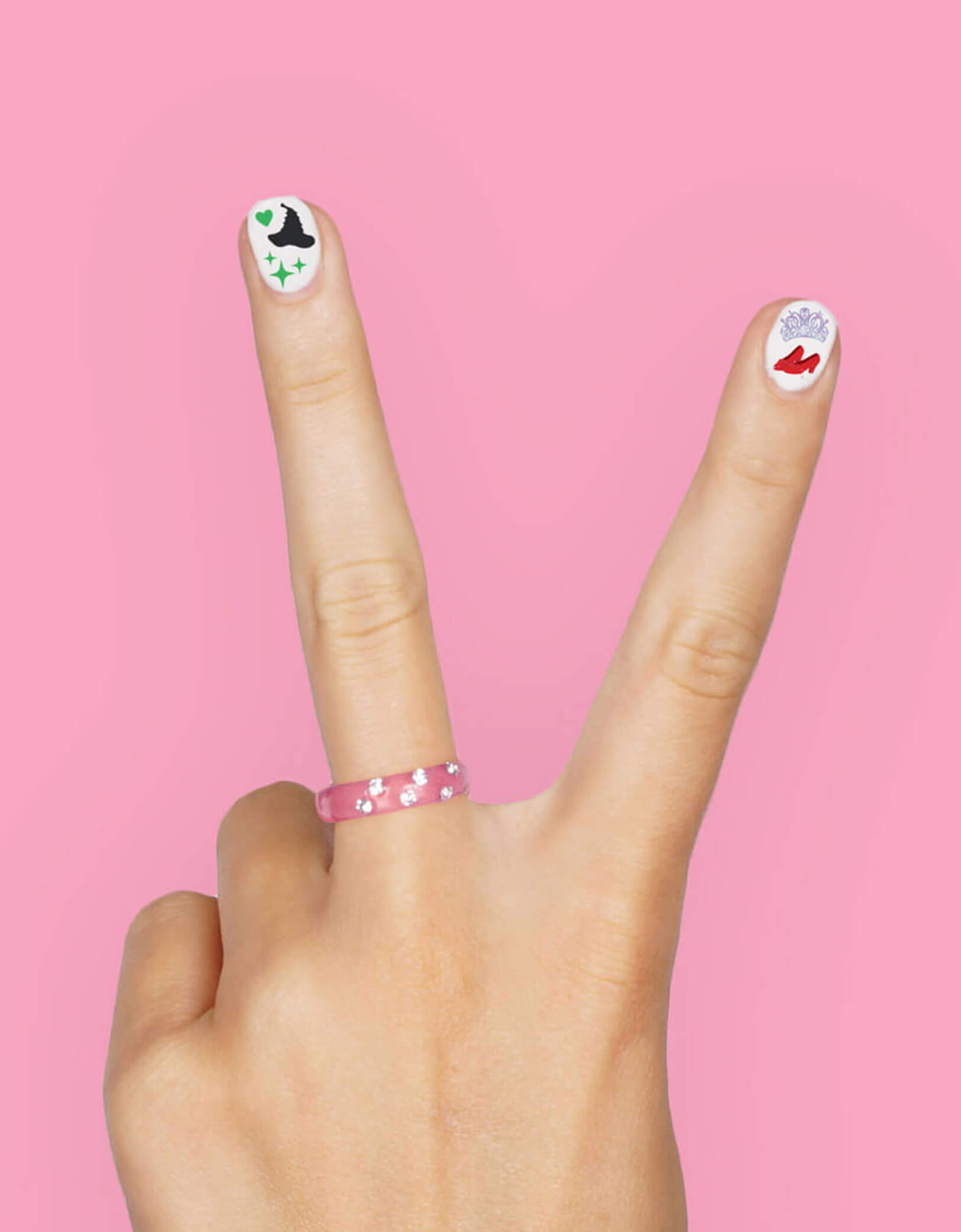 Hand making a peace sign with decorated Wicked themed nail stickers from Momo Party against a pink background.