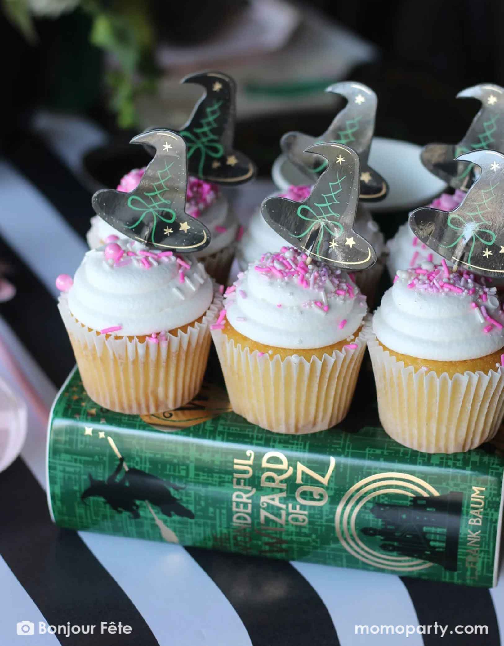 A few cupcakes with white frosting and pink sprinkles topped with black witch shaped toppers on a 'The Wonderful Wizard of Oz' book. A whimsical and magical party treat idea for kid's Wicked themed party.