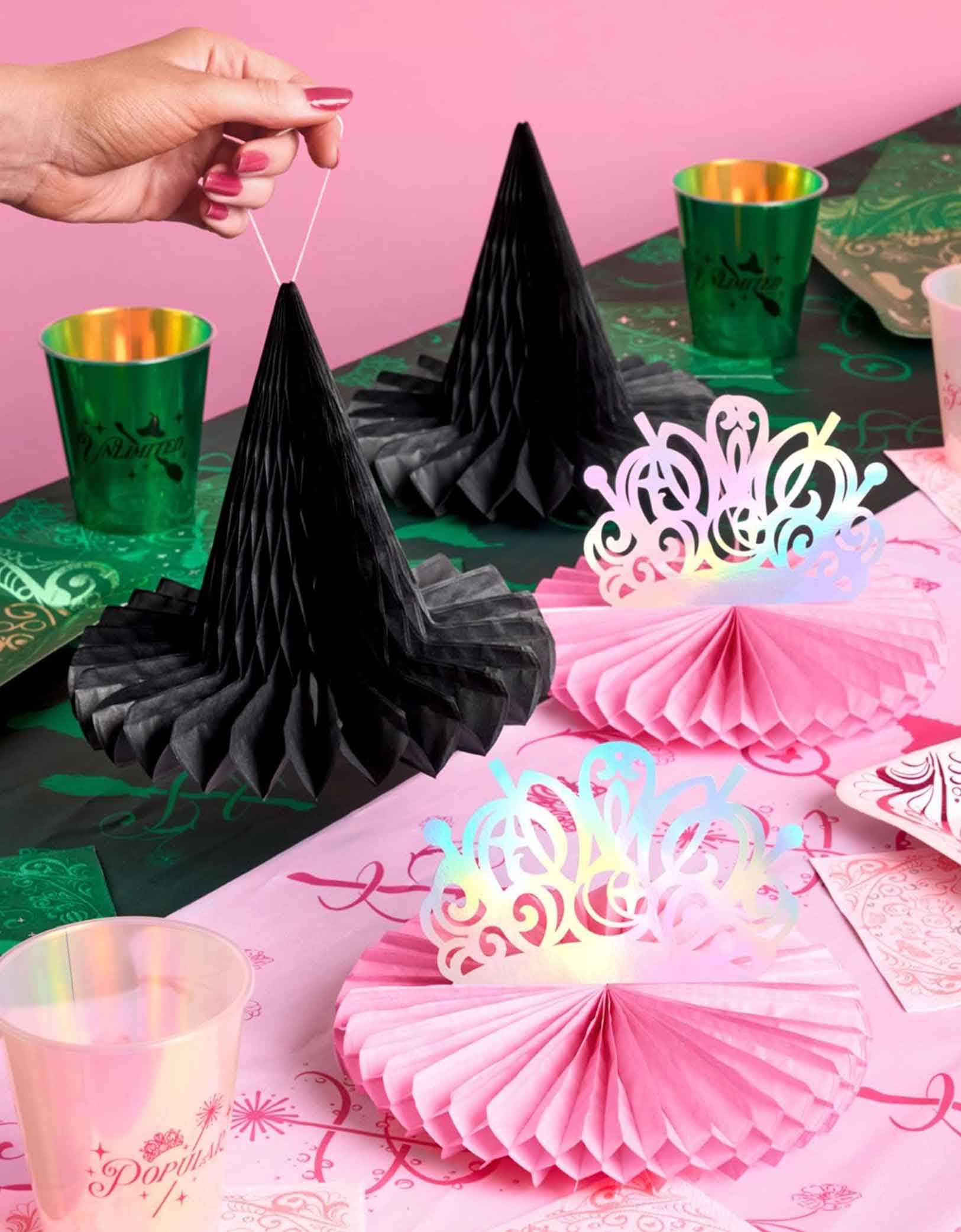 A festive Wicked themed party table featuring various of Wicked themed tablewares and decorations from Momo Party including Wicked Tiara & Witch Hat Honeycomb Set, Wicked party cups in green and green and Wicked party plates in green and pink on a Wicked themed tablecloth.