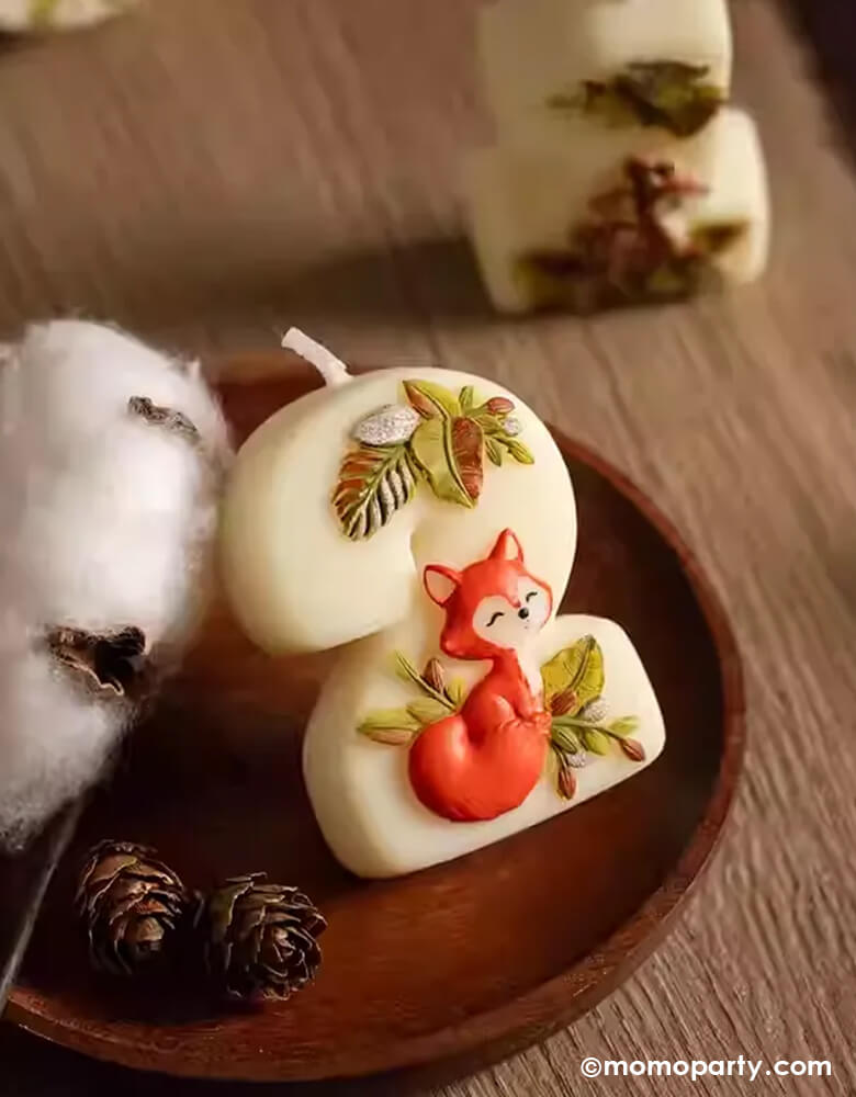 An adorable woodland themed kid's number two birthday candle a cute fox design and some greenery from Momo Party in a wooden tray filled with decorative cotton and pine cones filled with autumn and fall vibes. This charming candle is perfect for kid's woodland themed second birthday party celebration.