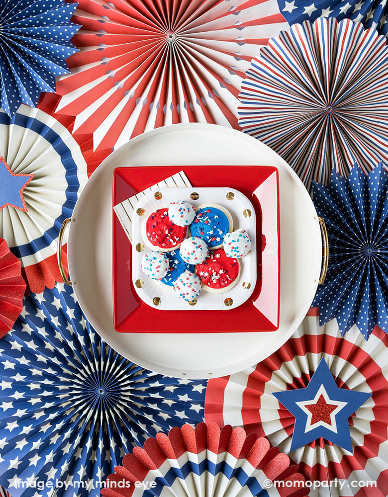 A top shot of 4th of July party table with Stacked Larger round white plate, with medium square red plates and small polka dots plates with blue and red cookies in the middle, my mind's eye Stars & Stripes Party Fans as background 