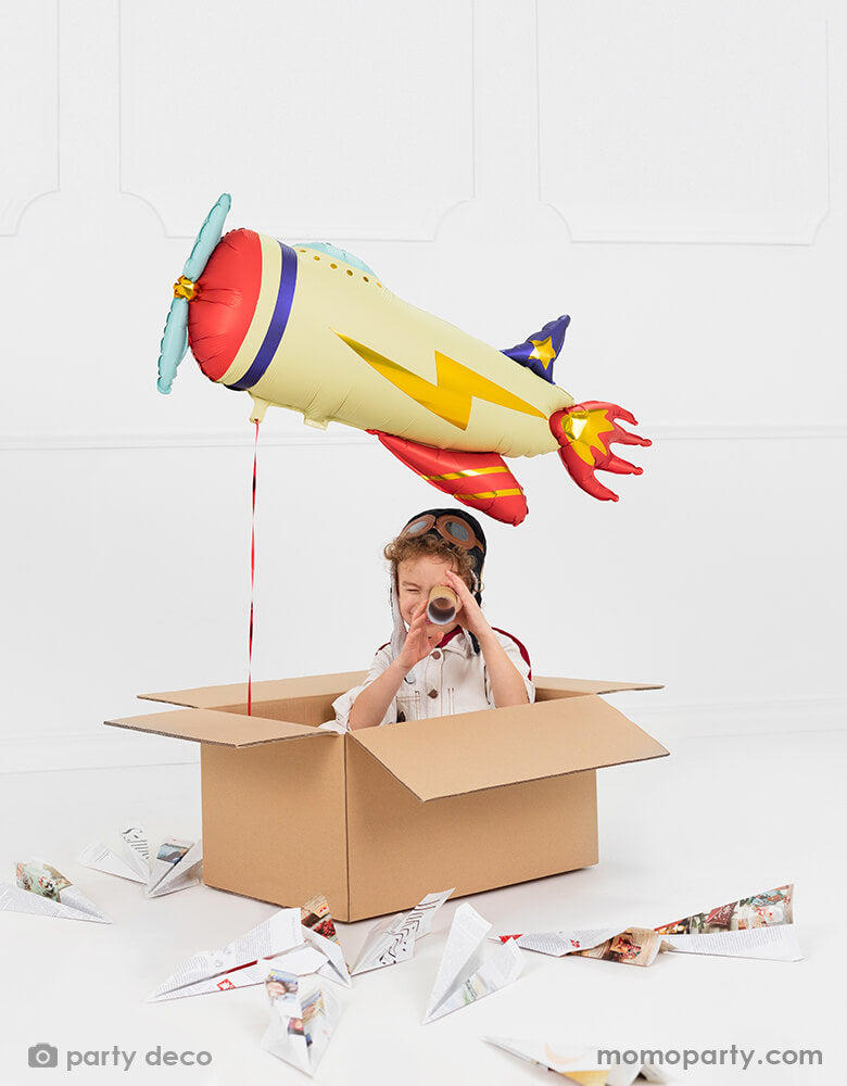 A little boy who dresses up as a pilot play make-believe in a Kraft shipping box surrounded with paper planes and with Party deco's 30" Party Deco 30" satin/matte vintage airplane shaped foil  balloon attached in the classic airplane colors of navy, red, blue and cream with lightning bolt gold foil design.
