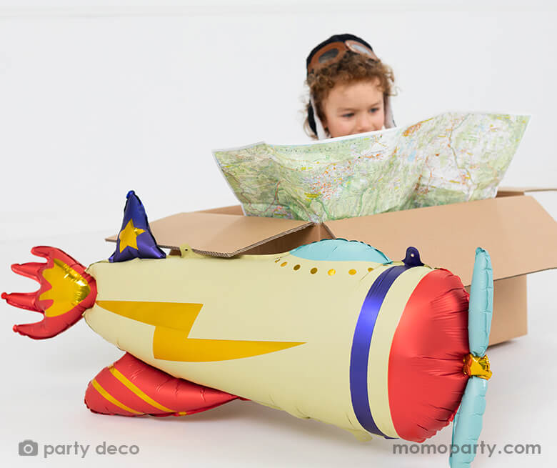 A little boy who dresses up as a pilot plays make-believe by reading its maps in a Kraft shipping box with Party deco's 30" Party Deco 30" satin/matte vintage airplane shaped foil balloon attached in the classic airplane colors of navy, red, blue and cream with lightning bolt gold foil design.