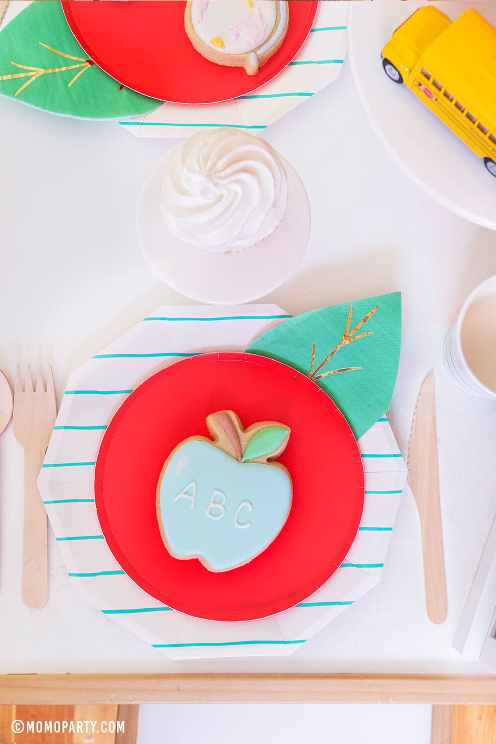 a Top view kid's table with a pastel ABC Apple cookie on top of Oh happy day Cherry Red side plate with Meri Meri Leaf napkin represent as Apple. Day Dream Society Aqua Striped Large Plates, wooden cutlery, cupcakes, school bus toys for a modern Back to school party celebration. 2020 Quarantine back to school home Party