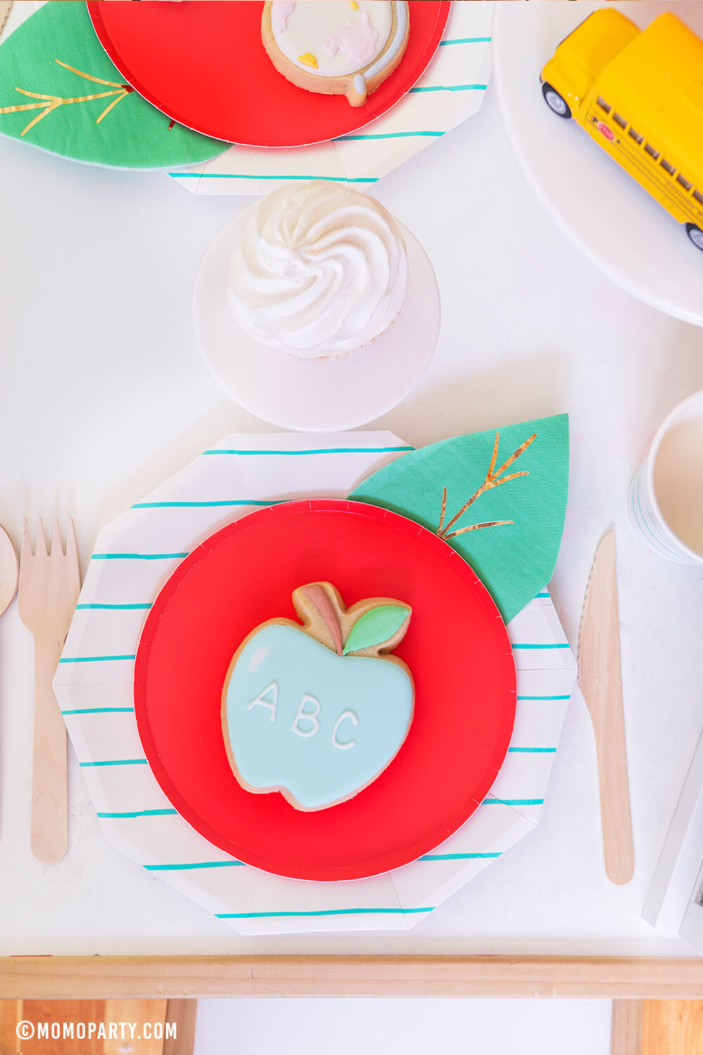 a Top view kid's table with a pastel ABC Apple cookie on top of Oh happy day Cherry Red side plate with Meri Meri Leaf napkin represent as Apple. Day Dream Society Aqua Striped Large Plates, wooden cutlery, cupcakes, school bus toys for a modern Back to school party celebration. 2020 Quarantine back to school home Party 