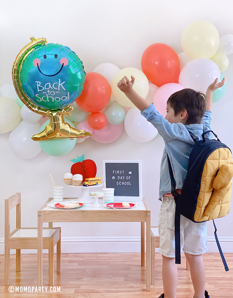 A Happy Boy wearing a backpack celebrate his Back to School party at home celebration with Anagram Back To School Globe Foil Mylar Balloon, pastel ballon garland, honeycomb apple, letter board with "first day of school", Mint stripe cups, plates...