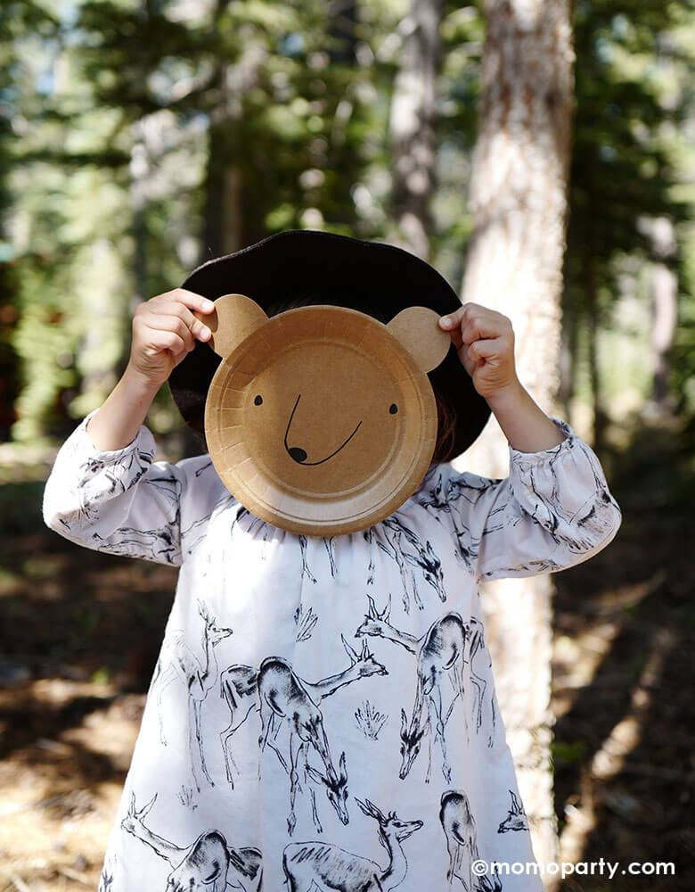 a girl wearing a white dress with deer illustration, holding Meri Meri bear plate as her face in a woods