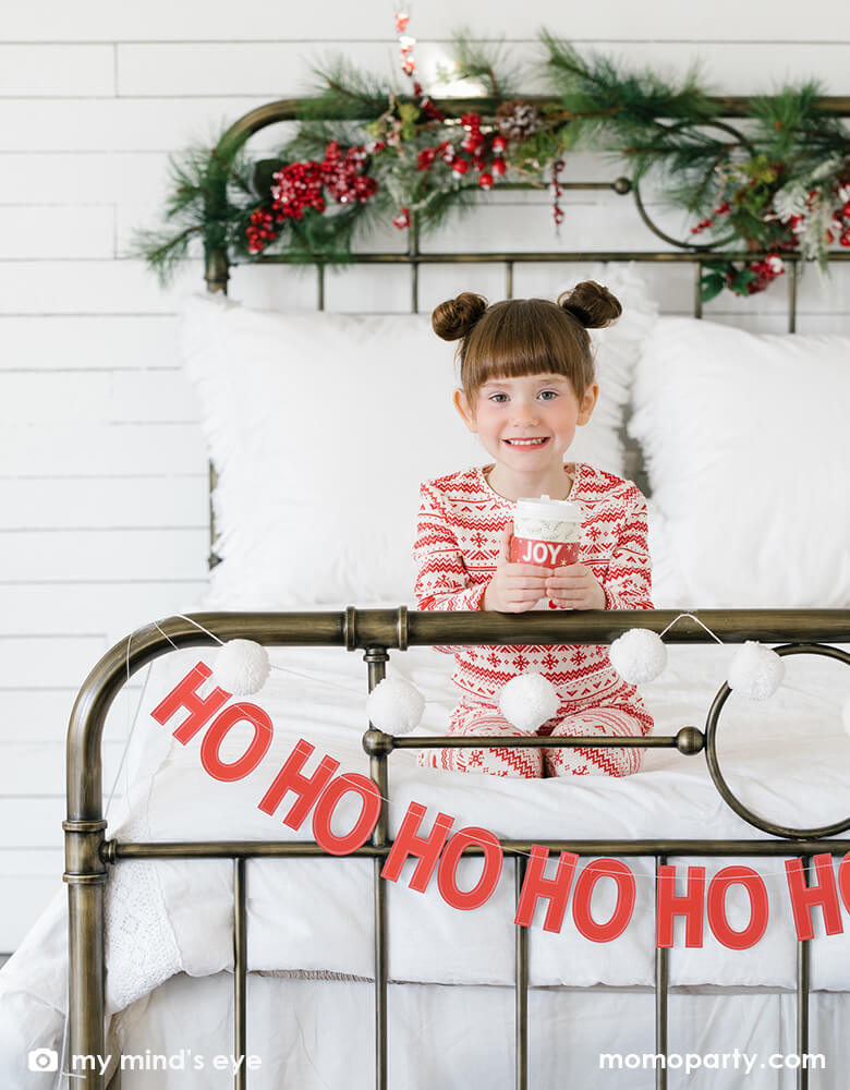 Holiday room decoration with My Mind's Eye - BEC804 Believe Ho Ho Ho Banner hanging on the end of the bed, with a kid wearing christmas red and white pajamas and holding a hot coco in the joy to go cup sitting on the bed. This 9 Banner pieces to complete the phrase "HO HO HO HO". Decorate for the Ho Ho Holidays this Christmas season with this jolly Santa Claus banner!