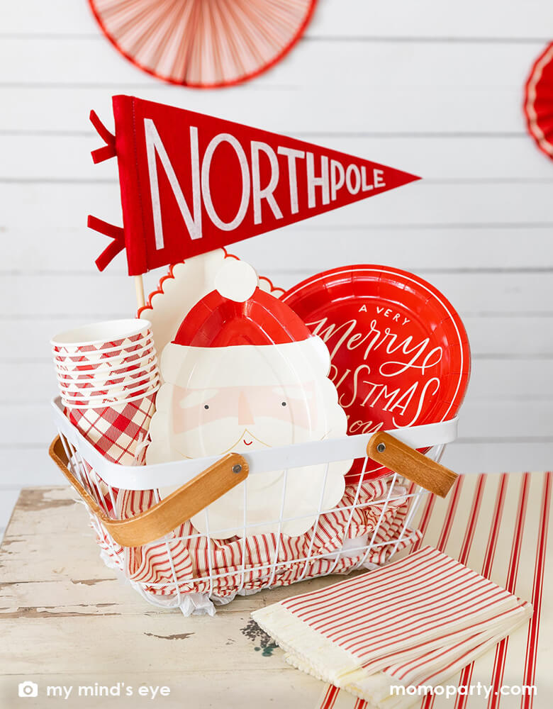 A basket filled with festive Christmas tableware in red from Momo Party including a Santa face shaped plate, some red checkered party cups, a scallop shaped dinner plates with red edge and a red round plates with words "A very Merry Christmas" on it, along with My Mind's Eye felt Northpole party pennant, a great idea for a family friendly Christmas party with kids