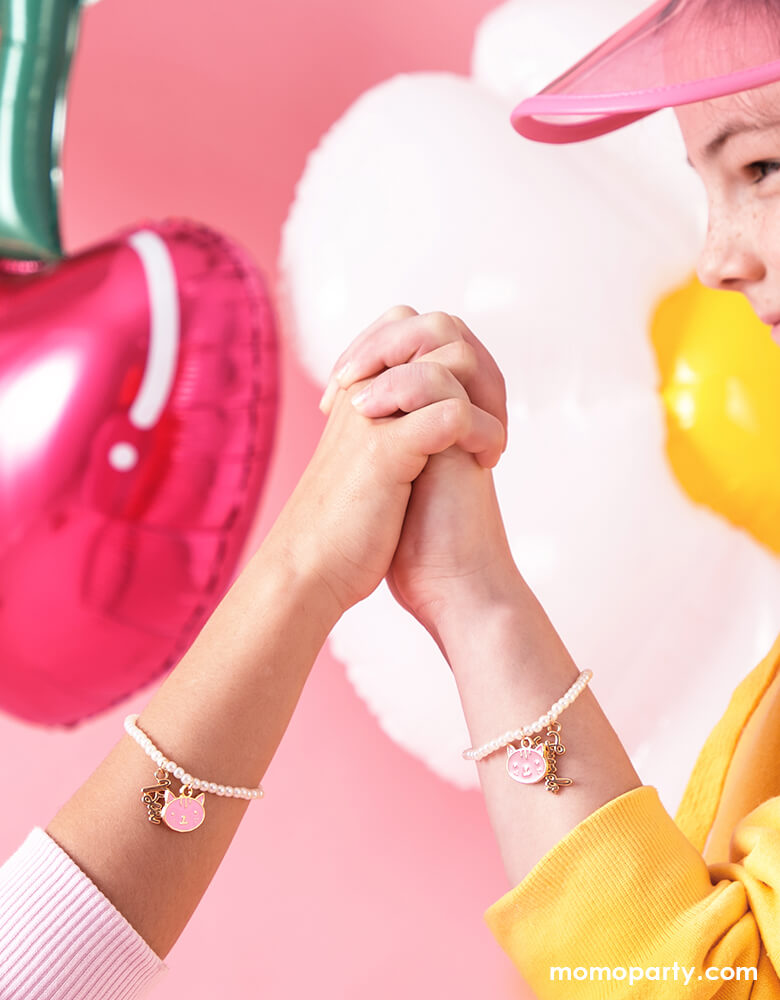 two girl best friends holding hands in front of a pink background featuring cute cherry shaped and daisy shaped foil balloons, both wearing Party Deco's Best Friends Bracelets, featuring two bracelets with pink kitty pendant and "best" "friend" word pendants. These adorable BFF bracelets are perfect for girl's Valentine's exchange!