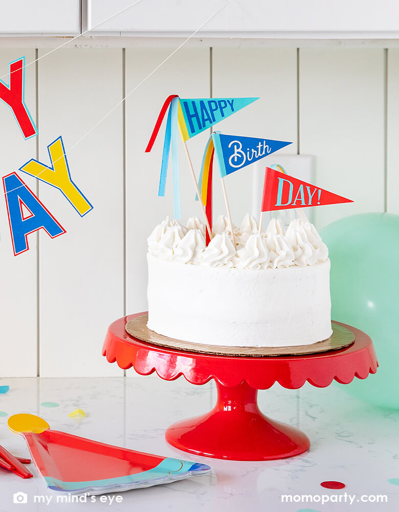 A cheerful buttercream birthday cake topped with My Mind's Eye Blue Birthday Cake Toppers in a set of 3 in cheerful colors of sky blue, navy and red, with message of Happy Birthday Day on each pennant with a background of festive and colorful birthday party balloons, perfect inspiration for a boy themed birthday party