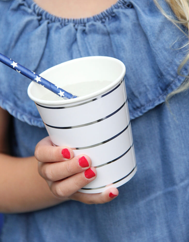 a Girl wearing a blue denim dress, and red nail polish, holding Day dream society Blue striped Party paper Cup with a blue stars paper straw in a 4th of july party celebration
