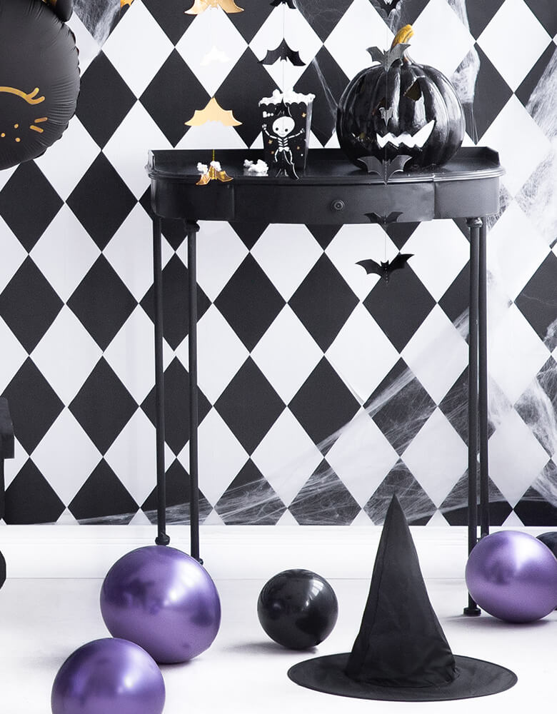 A party Deco Boo Treat Favor Box with Black Jack-o'-lantern decoration, bat garland on a black stand in front of a black and white diamond pattern wallpaper. A black witch hat on the floor with chrome purple latex balloons, black balloons for A modern black and white Halloween Party