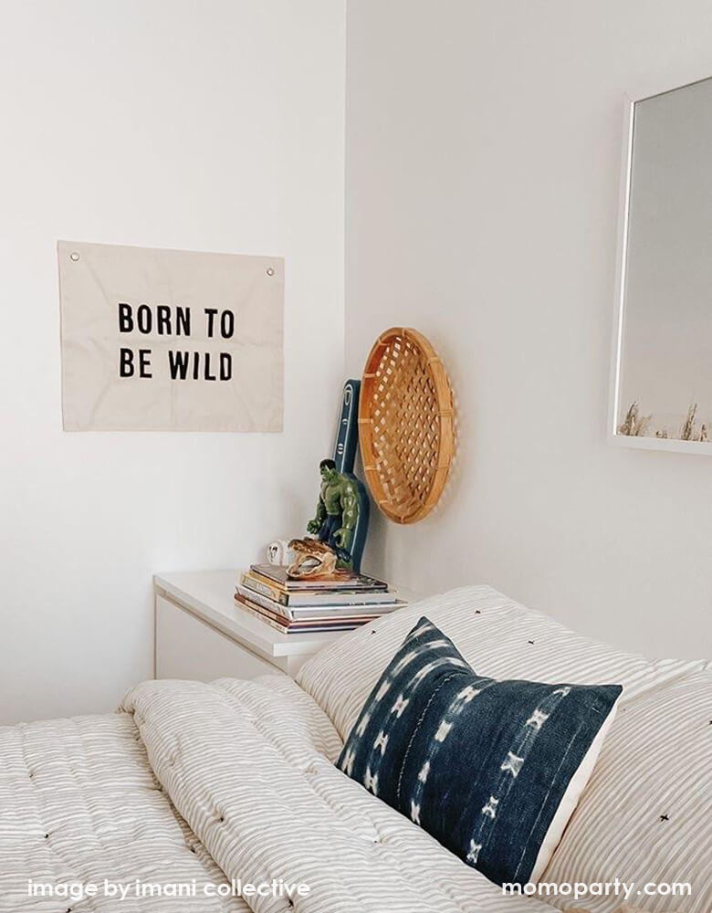 A California style bedroom, with a Imani Collective Born-To-Be-Wild-Banner, on top of a night stand with books, morden decorations. This modern hanging banner was sewn and screen printed by hand on natural canvas by local artisans in Kenya. It's a perfect decoration for your entrance hall, Nursery Decor, or your little wild one's playroom! Sold by Momo party store provided modern party supplies, boutique party supplies, chic holiday party supplies and high end party supplies
