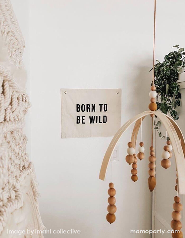 A California style bedroom, with a Imani Collective Born-To-Be-Wild-Banner, next to a modern gemestic mobile, green leaf plant, Woven Wall Hanging. This modern hanging banner was sewn and screen printed by hand on natural canvas by local artisans in Kenya. It's a perfect decoration for your entrance hall, Nursery Decor, or your little wild one's playroom! Sold by Momo party store provided modern party supplies, boutique party supplies, chic holiday party supplies and high end party supplies
