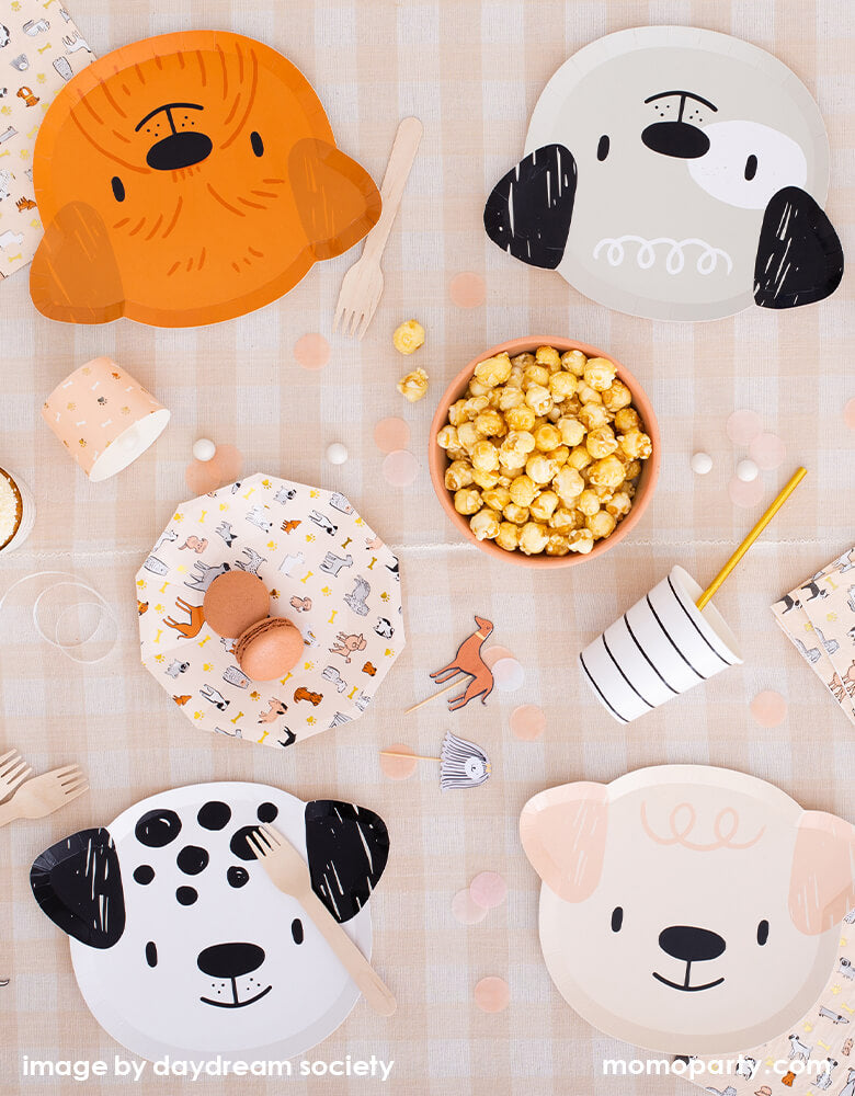 Bow Wow Dogs themed party table top view with Bow Wow Dog shaped Paper plates in 4 different dog design, cute dog toppers, popcorns in a bowl, pale brown colored macaron in the Bow Wow dog small plate, black striped paper cups with gold foil straw. wooden utensils, confettis on top of Natural Cabin Check Tablecloth. These warm neutral color themed modern Dog Birthday Party Supplies are perfect for a dog lover, a dog themed birthday party, Let's Pawty birthday party, adorable puppy themed party