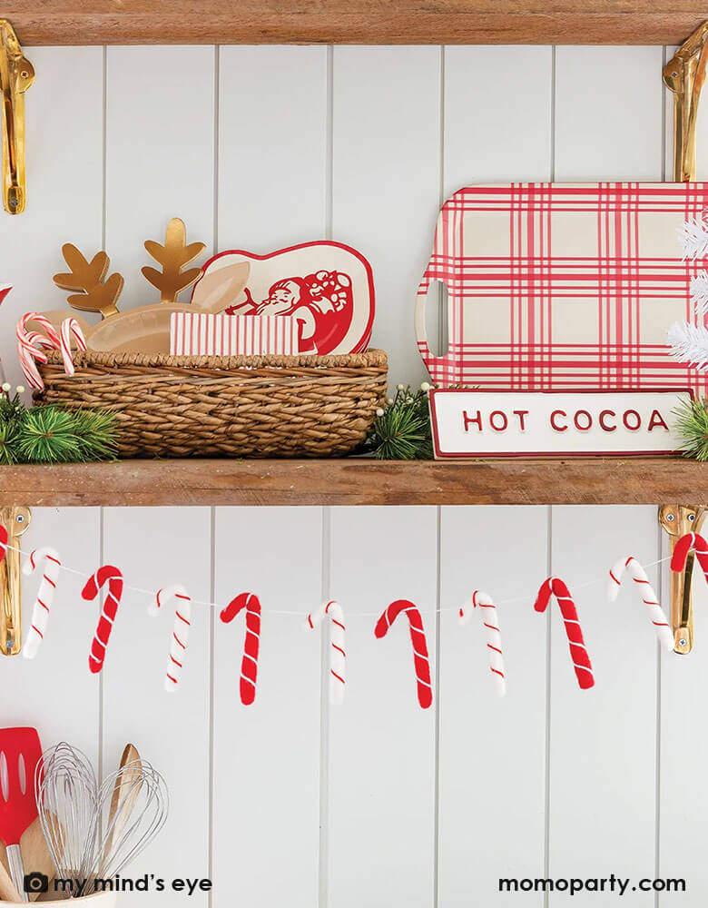 A kitchen counter shelf decorated with Christmas holly and momo Party's 7ft candy cane felt garland by My Mind's Eye, along side with festive partyware including a reindeer shaped plates, a set of paper plates with Vintage illustrated Santa, and a bamboo tray featuring red gingham design for hot cocoa, a perfect look and inspiration for this holiday season