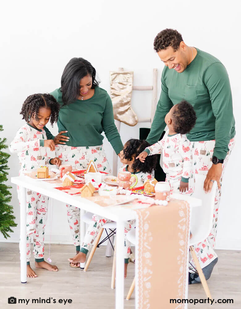 A family of 5 in their Holiday pajamas gathering at a table for a gingerbread house decorating activity featuring Momo Party's gingerbread house paper table runner draping over the table, on the tabletop is adorned with gingerbread house themed tableware including Momo Party's gingerbread house shaped plates, party napkins, gingerbread man mugs. Next to the table are some festive velvet stockings and a Christmas tree, ready for a fun kid's Holiday party!