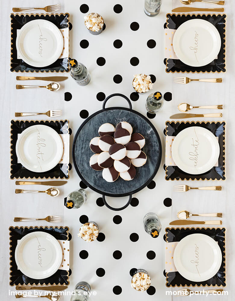 Top view of Modern celebration table top for 6 people, setting with my mind's eye Cheers small Plates layed with Black Scalloped Large Plates and gold polka dot napkins, with golden cutlery on the side, a glasses of drink, and sweets on a black tray on top of Cream with Black Dots Table Runner in the table center. These simple elegant partywares make your every celebration in style