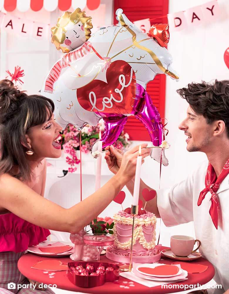 A loving couple at a dinner table enjoying a box of chocolate and pink cake to celebrate Valentine's Day. In the back the room is decorated with Momo Party's 32x34" Cupid shaped foil balloon and a bouquet of red heart shaped latex balloons, with a banner says Happy Valentine's Day in pink, it sets a romantic scene for a special Valentine's Day celebration.
