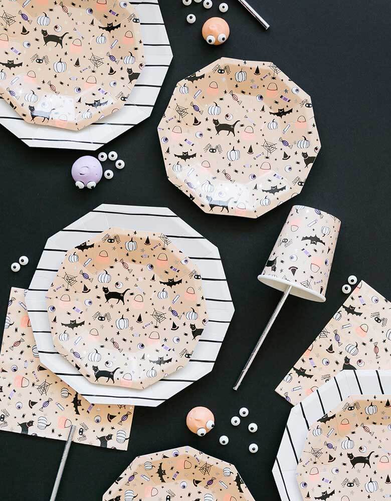 A spread of Daydream Society's Hocus Pocus party supplies including party paper plates, napkins, cups featuring cute illustrations of kid's favorite Halloween characters including black cats, witch hats, bats, spiders, pumpkins on a black background topped with Halloween googly eyes decorations