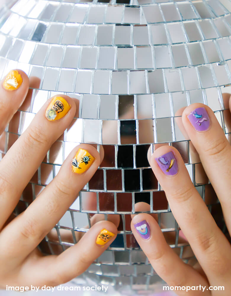 a kid hand holding a disco ball, she is wearing goldenrod color nail polish with Daydream society - into the wild nail stickers on her left hand fingers, and purple color nail polish with Daydream society - unicorns + rainbows nail stickers on her right hand fingers. These Safe and non-toxic nail stickers go on like pure magic and will have your nails looking great in no time!