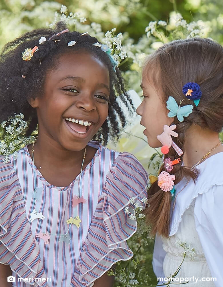 Two school aged girls chatting with each other wearing Meri Meri Easter themed accessories including bunny shaped felt hair clips, butterfly shaped felt hair chips and Meri Meri's acrylic leaping bunny necklace in 6 difference colors including  frosted blue, white, frosted pink, frosted mint, yellow and pink, with a gold tone chain and mint cord necklace.