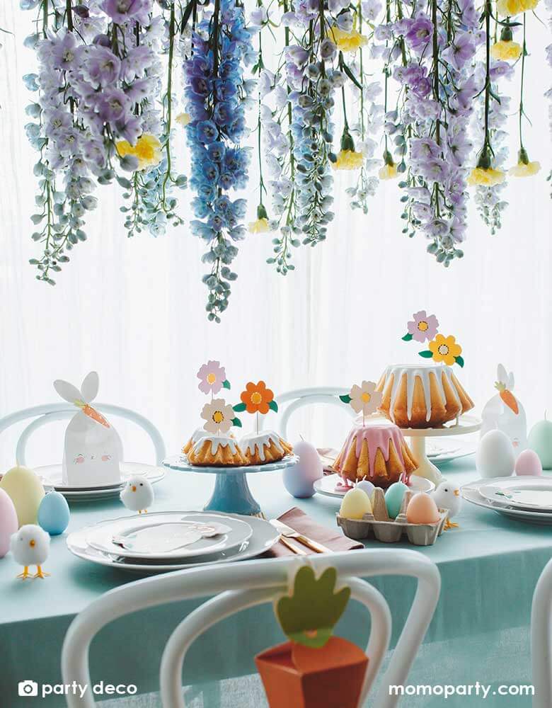 A beautiful table decorated with Easter themed tableware and decorations including Momo Party's 2.95 x 3.5 x 8.8" bunny treat bags by Party Deco, Easter chicks decorations, Easter egg shaped candles in pastel colors, and floral cupcake toppers on some bundt cakes. Above the Easter table there's wisteria flowers draping from above, makes a beautiful scene for an Easter celebration.