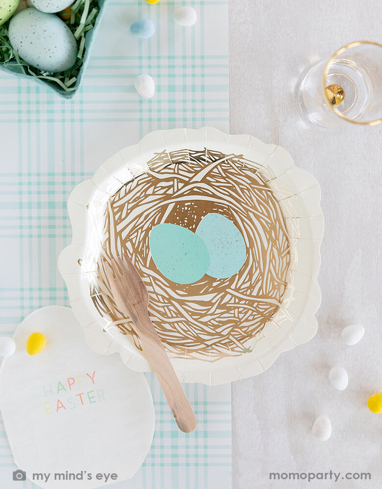 Top view of Easter party table top covered with My mind's eye -Nest shaped paper plates with Blue Plaid Guest Towel Paper Napkins and wooden fork, all on a blue checkered paper table runner, surrounded by colored eggs on mint shredded paper filling like a bird's nest and a glass of drink. These fresh partyware with modern fun designs are perfect for Easter celebrations