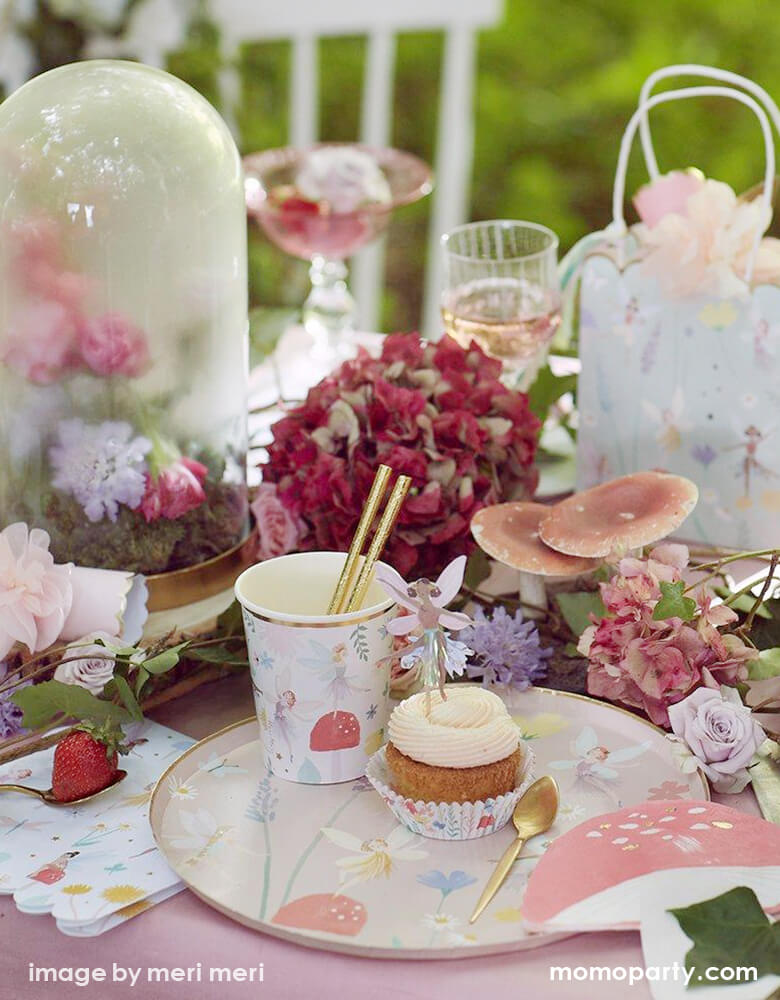 A outdoor Fairy themed birthday party table close up look of a cupcake with Fairy cupcake topper, and a fairy paper party cups with a gold foil straw on top of a Meri MerFairy Dinner Plate,  lots of flowers, mushrooms, leaves, strawberries around as decoration, such a dreamy modern look fairy party for kids, Tea party party with mom and daughter, perfect idea for a girl's birthday party, garden party, spring party, summer party