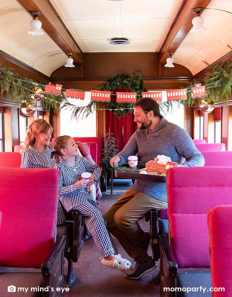 A family of a dad and two little girls in their Holiday pajamas enjoying a vintage tin tray filled with holly branches, holiday treats and cookies and Momo Party's North Pole Express train shaped plates, bell shaped side plates and polar express red 8 oz to-go cups by My Mind's Eye sitting on a vintage Polar Express train cabin decorated with Momo Party's North Pole Express train and bell garland set, a perfect idea for this Holiday season's celebration with family and kids.