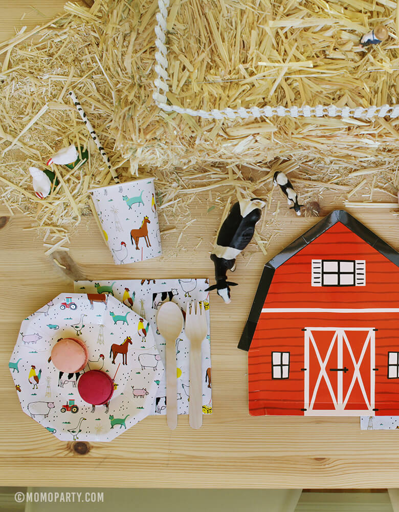 Farm Barnyard Party table inspiration set up with Macarons on the Farm-Animals Small Plates, wooden utensils on top of Farm-Animals Napkins, Cow pattern paper straw inside of Farm animal party Paper Cups, Red Barn house plate, animal toys on the Straw Bale as center piece
