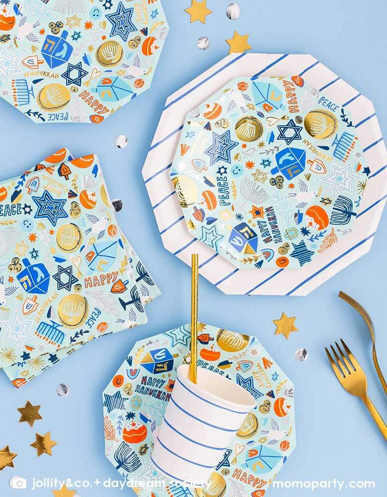 A tableware arrangement of Momo Party's 6.5" Festival of Lights Large Napkins and side plates by Daydream Society featuring Hanukkah themed symbol including dreidels, star of David, nine-branched candelabra, Menorah candles and Sufganiyah in blue colors with gold and silver foil detail, along with blue striped party cups and large plates, all together makes a modern Hanukkah tablescape
