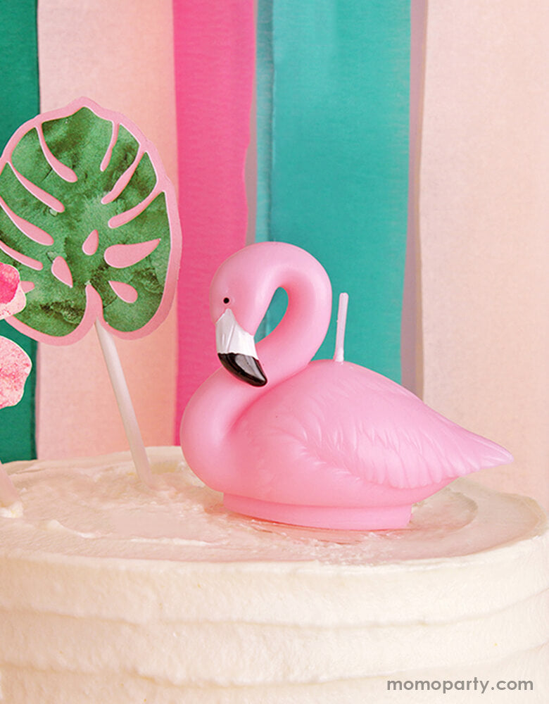A flamingo shaped birthday candle and a palm leaf shaped candle from Momo Party sitting on a buttercream white cake. Behind the cake is some colorful tropical themed paper streamer garland. Making this a modern and chic inspiration for a tropical themed party in the summer time!