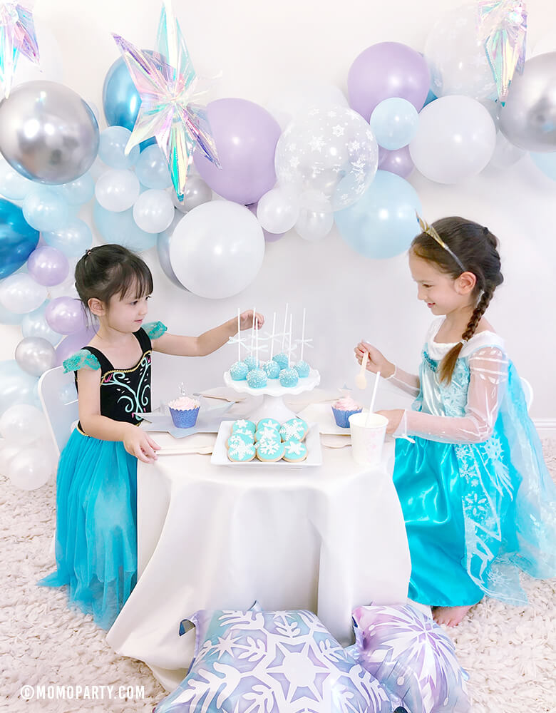 2 girls wearing Disney frozen frozen princess elsa and anna's costumes, sitting on the desert table, with a beautiful balloon garland with Pearl purple, Chrome blue, pearl white, pearl blue, clear snowflake latex balloons, iridescent hanging starts back drop,  celebrating a Girl's Frozen birthday party