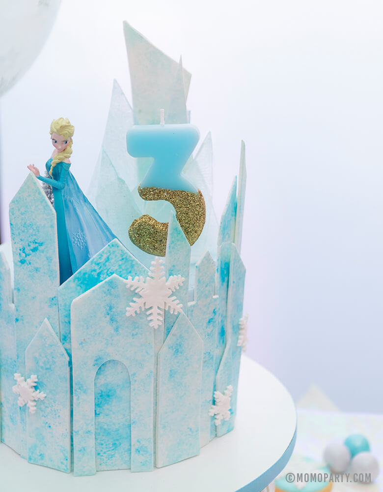 a Close up look of a Frozen Themed icy castle cake with Elsa Princess figure toy as a cake topper,  Talking Tables - We Heart Birthday Glitter Number 3 Candle in Blue and gold glitter, for a 3 years girl's disney frozen themed birthday party